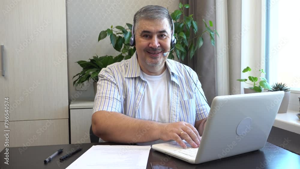 A man with a headset works at home on a computer. He looks at the screen then turns and smiles at the camera. Suitable for content about freelancing or remote work.
A man with a headset works at home on a computer. He looks at the screen then turns and smiles at the camera. Suitable for content about freelancing or remote work.