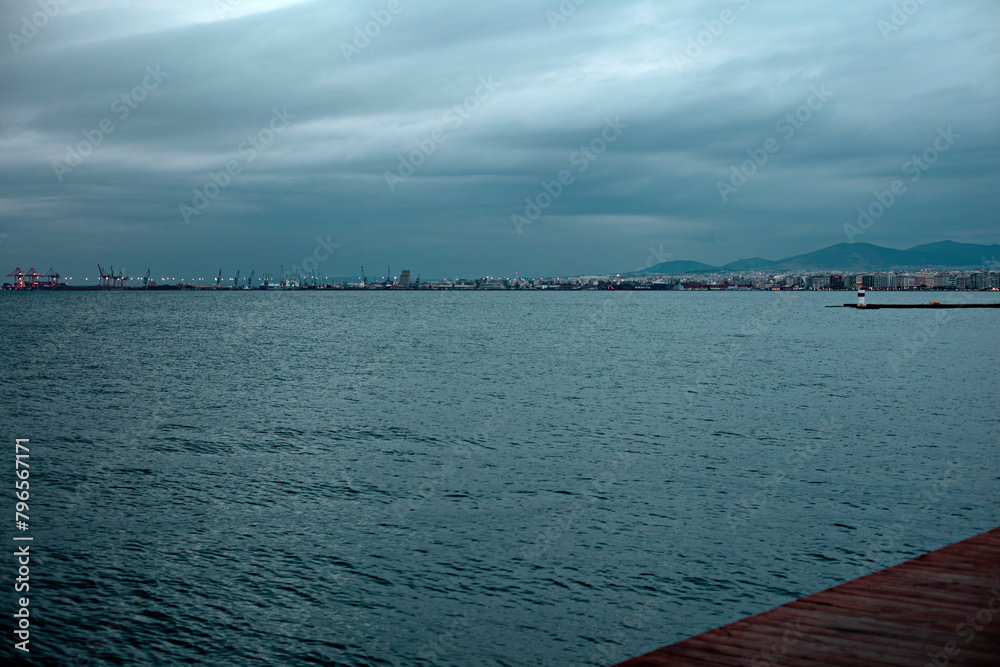 Dark clouds loom above the sea near an urban shoreline, with a hint of cityscape in the distance.
