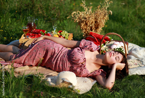 Young beautiful woman with short hair in pink dress having picnic in park on summer evening. Romantic leisure time activity