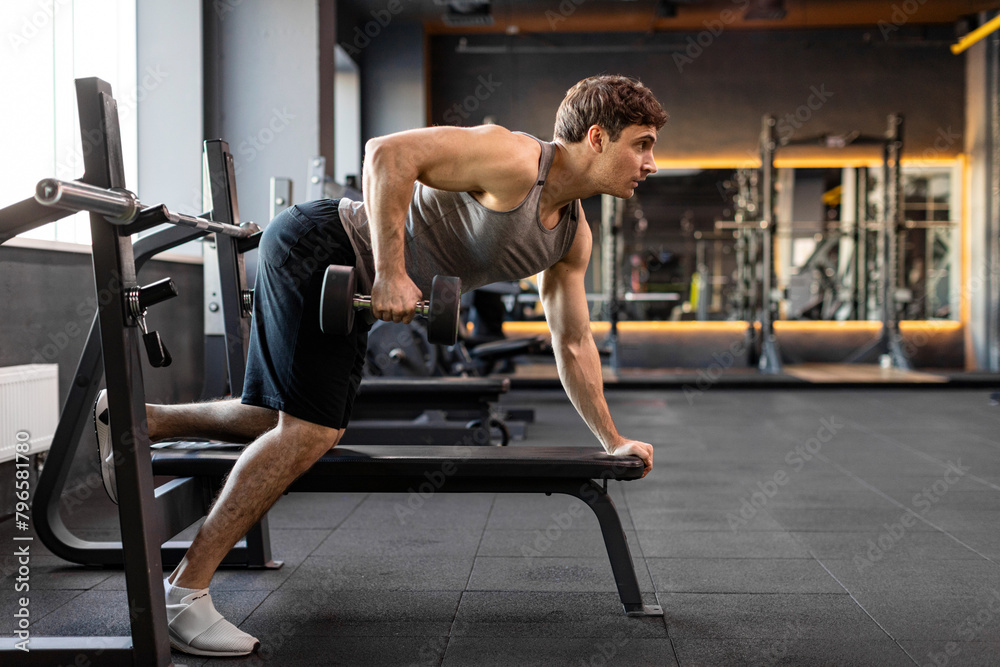Muscular bodybuilder man doing exercises with weights, lifting dumbbells at bench in gym interior, side view, free space Muscular bodybuilder man doing exercises with weights, lifting dumbbells at bench in gym interior, side view, free space