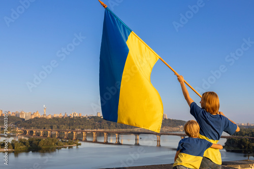woman and child with yellow-blue flag stand high on the roof of a house against of sky and Dnieper River in Kyiv. Patriotism, drawing attention to war in Ukraine. Support Ukraine. Independence Day