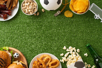 Football party setup. Overhead view of game day snacks: chips, pistachios, popcorn, onion rings, beer, soccer ball, referee gear, and more on grass background with space for text