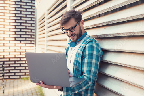 Happy hipster guy using his laptop computer while standing outdoors on a sunny day