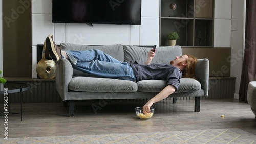 A young red-haired guy is lying on the sofa and eating corn flakes, scrolling through social networks on his phone photo