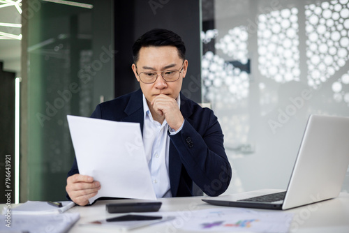 A man in a suit is reading a piece of paper while sitting at a desk with a laptop. He is deep in thought, possibly contemplating a business decision or a personal matter