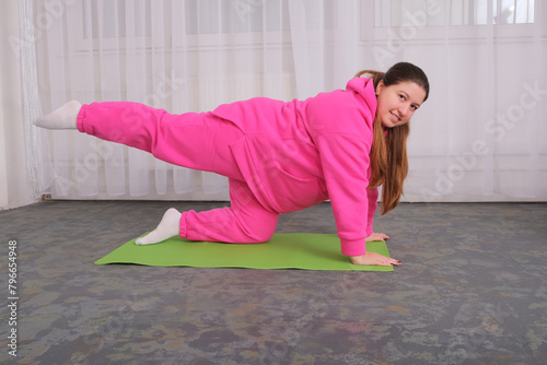 An overweight girl in a pink tracksuit is doing sports on a fitness mat