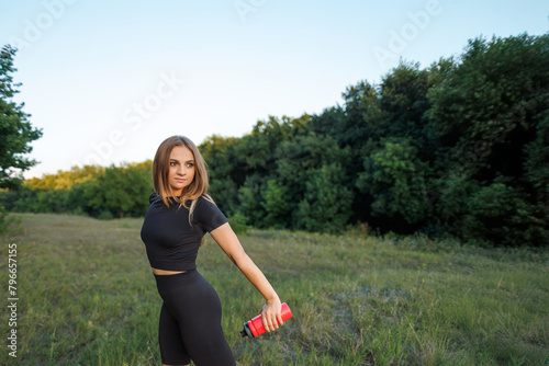 The athletic girl stands sideways and holds a red water bottle in her hand. Athletic girl in a tight uniform working outdoors in the park.