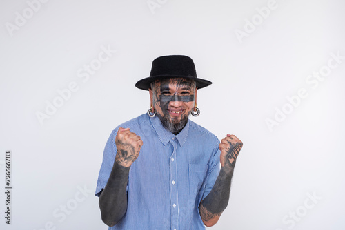 Fashionable Asian man with face and eye tattoos, including flesh tunnel and nose and lip piercings, elatedly fist pumps against a white background. photo