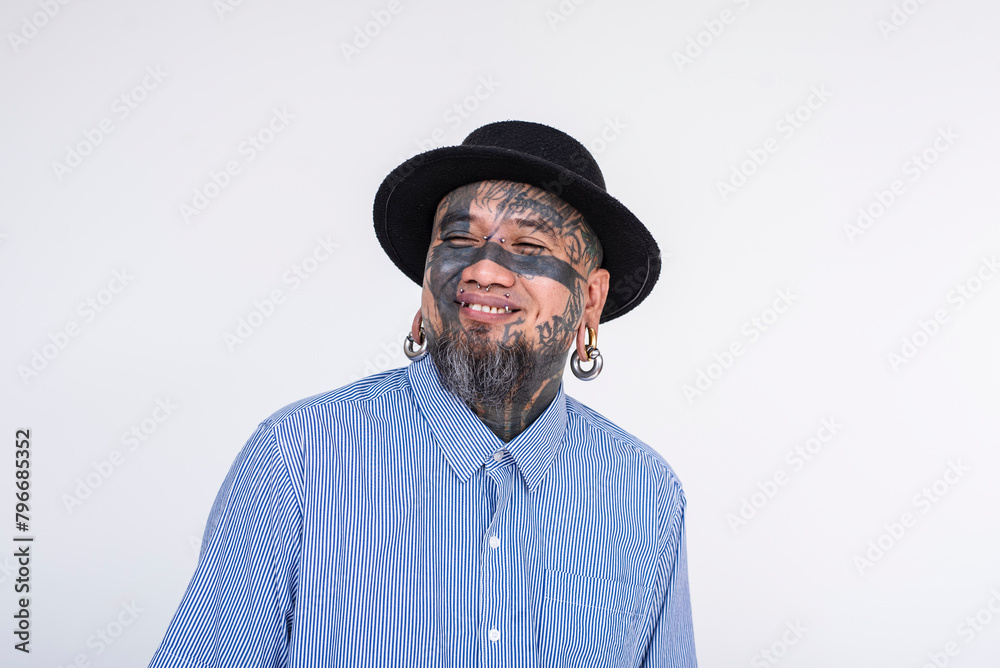 Portrait of a cheerful Asian man fully covered in facial tattoos, showcasing eye tattoos and diverse body piercings, dressed in a stylish hat and striped shirt against a white backdrop.