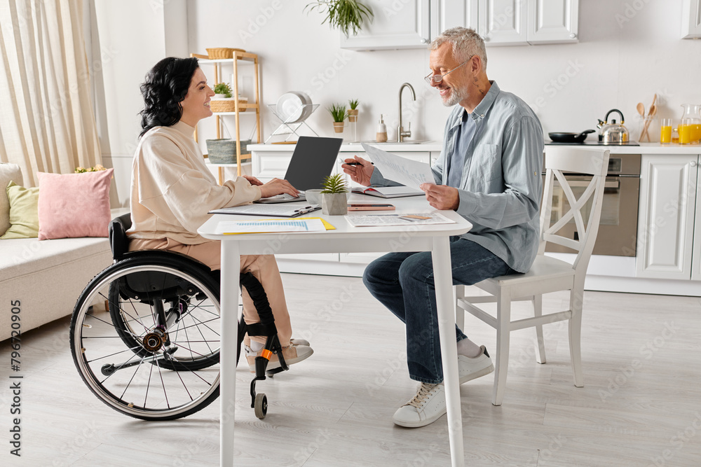 A man and a woman in wheelchair sit together at a table in their kitchen at home. A man and a woman in wheelchair sit together at a table in their kitchen at home.
