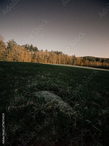 A large stone in the middle of a farm field. Hay is grown in the field that is illuminated by a full moon. Stars are visible above the forest. The constellations perseus, cassiopeia and cepheus  seen. photo