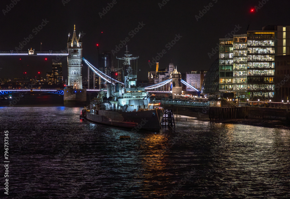 Front view of the war museum ship HMS Belfast docked on the River Thames with reflections of lights from restaurants and buildings in the water in front of Tower Bridge illuminated at night.