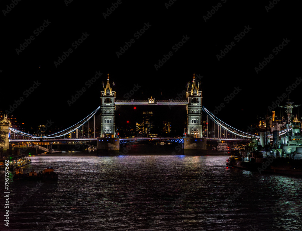 Obraz premium London bus driving past Tower Bridge illuminated at night with illuminated skyscrapers and the war museum ship HMS Belfast docked on the River Thames with reflections of restaurant lights in the water