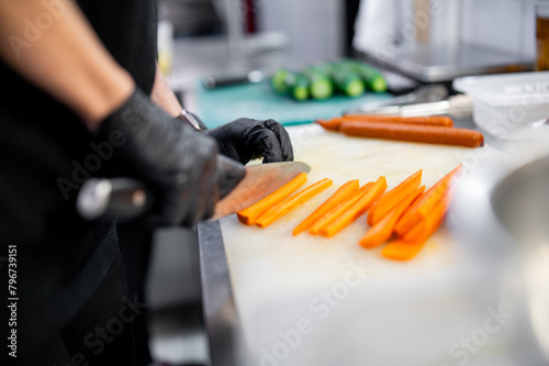 Skilled chef in black attire meticulously slicing fresh carrots on a white chopping board in a well-equipped kitchen.