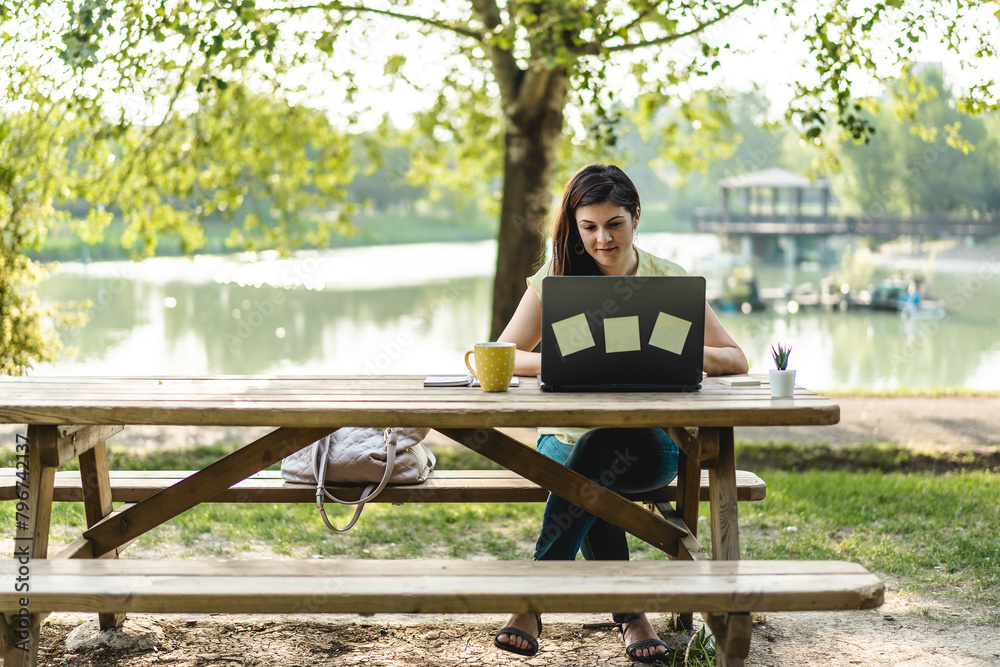 Fototapeta premium Freelancer working in nature's calm. Serene park setting enhances concentration and well-being. Woman at remote work.
