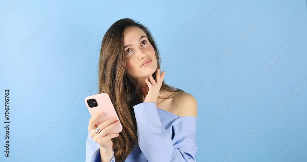 Calm beautiful young woman isolated on blue background. Girl in blue pullover flirting while chatting on mobile phone, dreaming. Woman think about answer, touch lip, look at side, unsure. Calm beautiful young woman isolated on blue background. Girl in blue pullover flirting while chatting on mobile phone, dreaming. Woman think about answer, touch lip, look at side, unsure.