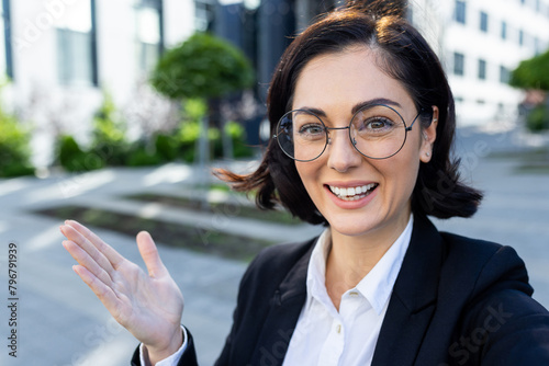 Smiling young woman in business suit standing outside and talking on video call on phone, gesturing with hands. Close-up photo