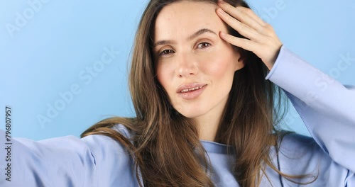 Brunette close up girl stares at the phone and fixes her hair. Female uses phone like mirror, peace gesture, blow kiss. Young girl make photo from hands on blue background, look at camera and smiling. photo