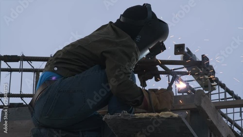 Worker performs welding work in protective mask is weld metal and is sitting at an altitude against sky background. Close up.