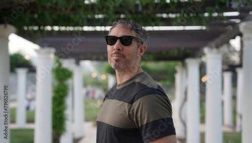 Portrait of a man in his 30s, wearing sunglasses in the park at sunset. The white pergola columns in the background. 