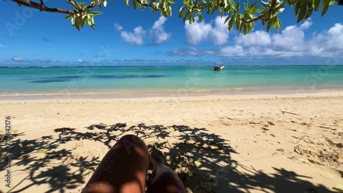 Women's legs with their feet stretched out on the white sand in the shade of the local and palm trees of the topical island. Opposite the turquoise blue ocean and vast sea. (ID: 796842176)