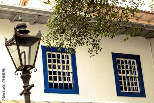 Poste de ferro, janela de madeira de cor azul, plantas no telhado e parede branca.  photo