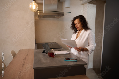 Beautiful Latin American woman reads paper bills pay online at home. Pretty brunette holding documents and bank letter, managing account finances, calculating taxes, planning loan debt payment