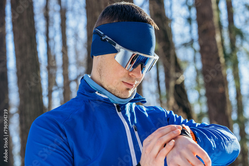 Focused male skier in a blue outfit and reflective goggles checks his fitness tracker amidst a snowy woodland environment.