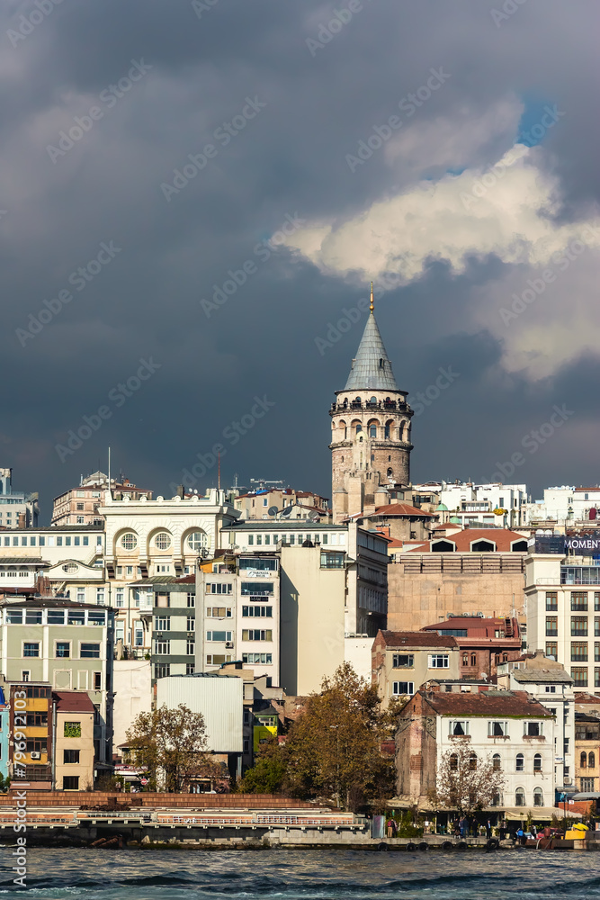 The Galata Tower stands tall amidst the modern Istanbul skyline under a dramatic cloud-filled sky. Istanbul, Turkey (Turkiye)