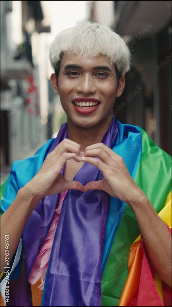 Transgender Individual with Makeup Wrapped in Rainbow Flag, Showing with Hand Heart Gesture Outside. Fashionable Genderqueer Person Standing Outdoors, Displaying a Heart Sign for the Camera Transgender Individual with Makeup Wrapped in Rainbow Flag, Showing with Hand Heart Gesture Outside. Fashionable Genderqueer Person Standing Outdoors, Displaying a Heart Sign for the Camera