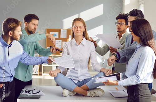 Calm, tranquil woman sitting on office table in lotus yoga pose, mediating, chilling out, and ignoring team of diverse colleagues who are demanding to do lots of work as soon as possible photo