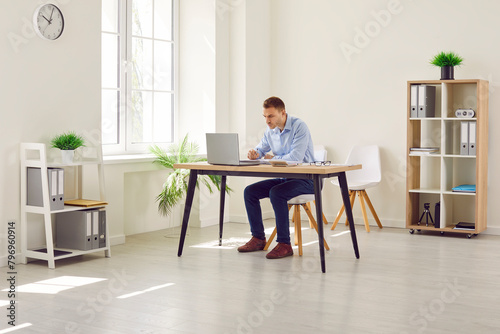 Full lenght portrait of a confident attractive young business man working on a laptop in modern office. Company employee sitting at the desk of his workplace making notes and finance calculations. © Studio Romantic