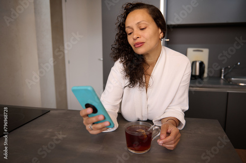 Attractive Latin American woman using smartphone while drinking morning tea in kitchen at home. Relaxed positive lady reading text messages on her mobile phone and smiling