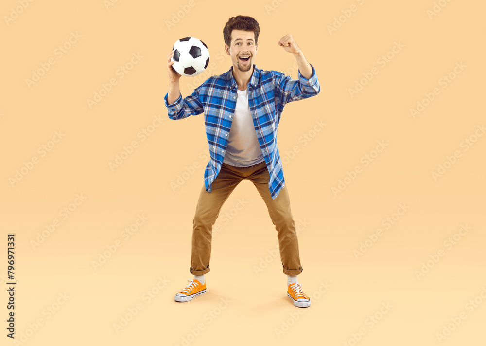 Cheerful excited man supporting his favorite soccer team during world cup match. Guy with funny facial expression holding soccer ball and clenching fist on beige background. Concept of football fans.
