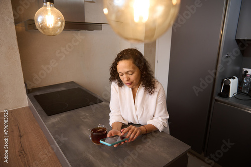 Gorgeous young Latin American brunette using mobile phone for online communication. Happy Arab woman browsing social networks on smartphone and drinking tea in the kitchen at home. Free space for ads