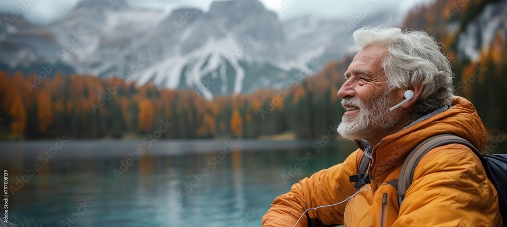 A senior man, with white hair and a trim beard, sits by a lake with mountains behind him, smiling as he enjoys music through earbuds. A senior man, with white hair and a trim beard, sits by a lake with mountains behind him, smiling as he enjoys music through earbuds.
