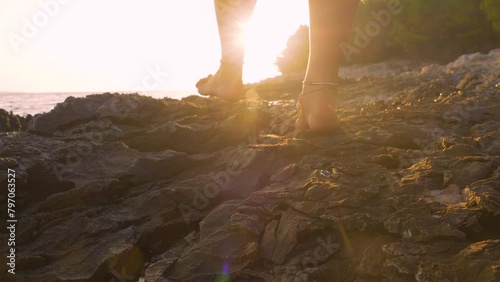 LENS FLARE, CLOSE UP: Barefoot lady and a dog walk carefully on sharp coastal rocks of rugged Adriatic beach. While they stroll along rocky seashore, golden summer sun is slowly setting behind horizon photo