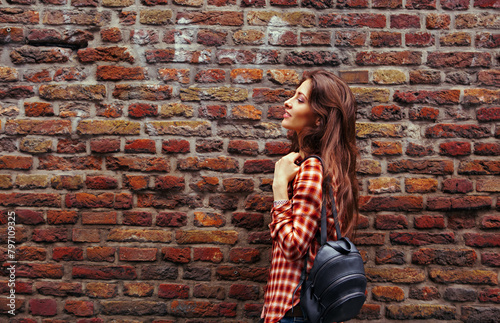 Beautiful brunette smiling woman with long brown hair standing on the old red brick wall building in casual red shirt holding shoulder leather black bag on the spring city. Style