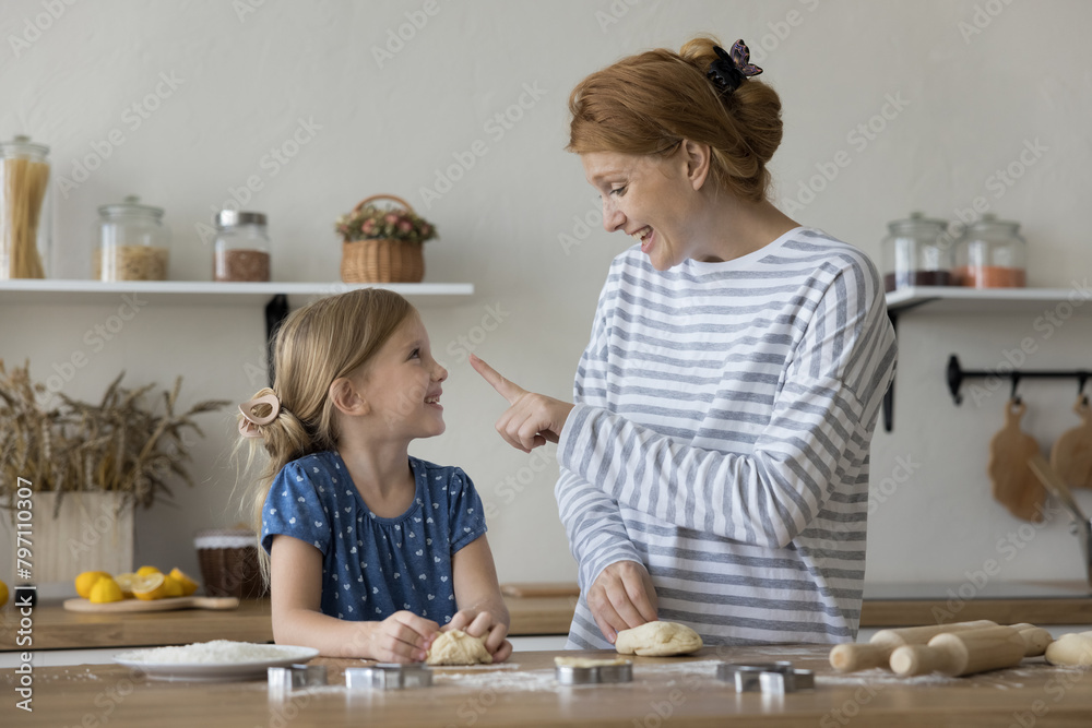 Naklejka premium Little beautiful girl helper cooking with loving young mother, kneading dough, preparing sweet homemade cookies on holiday in cozy kitchen. Parent teach kid to cook, sharing traditional family recipe
