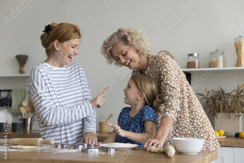 Little 6s girl cooking with loving mom and granny in kitchen, playful multi-generational family having fun, laughing, enjoy cookery and communication on weekend at home. Ties, culinary, joint hobby © fizkes