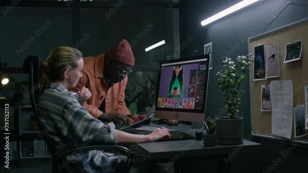 Medium side of Caucasian female graphic designer working on commercial images on computer in postproduction studio, African American male colleague showing reference on tablet, giving recommendations Medium side of Caucasian female graphic designer working on commercial images on computer in postproduction studio, African American male colleague showing reference on tablet, giving recommendations