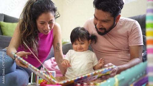 Happy hispanic family reading a learning book to their baby daughter, young latin couple enjoying time with their cheerful happy toddler