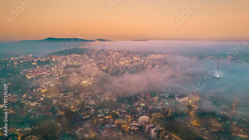 An aerial view of a foggy city in the morning. The fog is white and thick, and it covers most of the buildings. The city is built on hills, and there are trees on the slopes. The sky is clear blue.