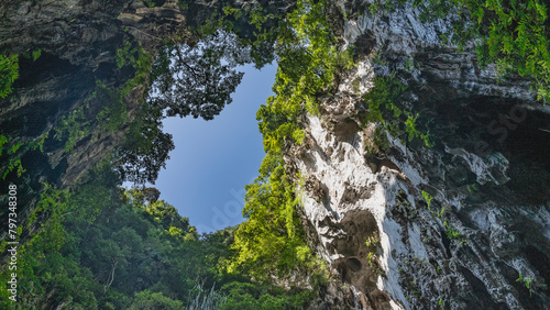 A bright blue sky surrounded by a frame of sheer cliffs. Green tropical vegetation grows on steep limestone slopes. A hole in the ceiling of the cave. Malaysia. Kuala Lumpur. Batu caves