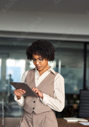 Young busy professional African American business woman company executive or entrepreneur using digital tablet working standing in office looking at pad, managing financial project data. Vertical.