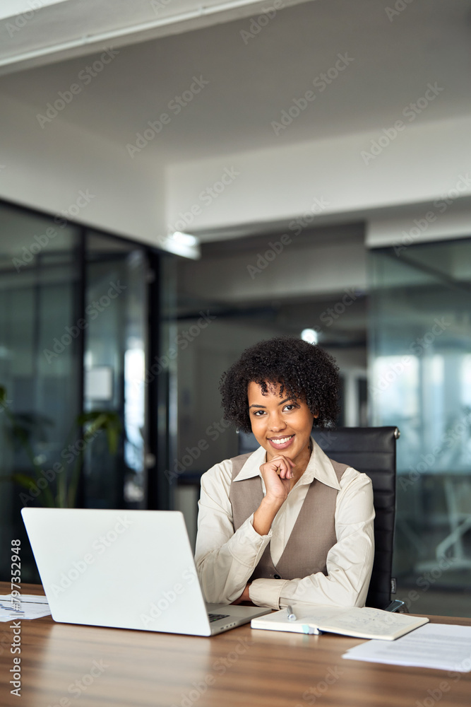 Confident happy young female lawyer, smiling professional African American business woman company manager executive working on laptop in office sitting at desk looking at camera, vertical portrait. Confident happy young female lawyer, smiling professional African American business woman company manager executive working on laptop in office sitting at desk looking at camera, vertical portrait.