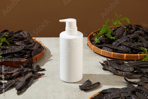 High angle shot photo with unlabeled white bottle stand isolated on light gray table against on brown background with some locust fruit scattered lay around. Mockup for designing with space for text photo