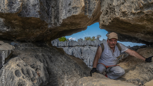 A tired man with a backpack and safety equipment crouched in the opening of the rocks, leaning on his hands. Behind, against the blue sky, steep karst cliffs with sharp peaks are visible. Madagascar. photo