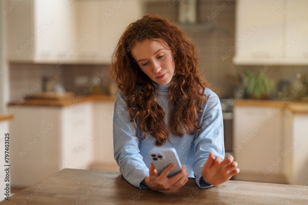 unhappy redhead woman sitting on sofa, using mobile phone, talking, surfing internet, reading, watching videos in kitchen at home. Toned image unhappy redhead woman sitting on sofa, using mobile phone, talking, surfing internet, reading, watching videos in kitchen at home. Toned image