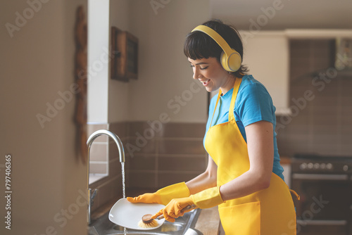 happy woman washing dishes in kitchen and listening to music with wireless headphones,  enjoying doing domestic chores